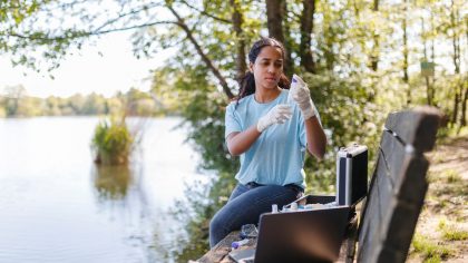 Jeune femme portant des gants réalise des recherches environnementales près d'un lac. Elle utilise du matériel, y compris un ordinateur portable et des kits de test. Des arbres et de l'eau en arrière-plan.