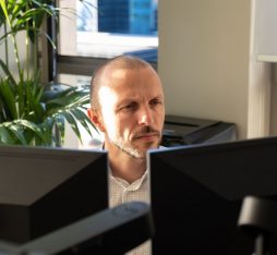 The image shows a man sitting at a desk, focused on two computer screens in front of him. He has short hair and a well-groomed beard. He is wearing a white shirt with subtle patterns. In the background, there is a green plant that adds a touch of nature to the work environment. Natural light is coming through a window, illuminating the scene.