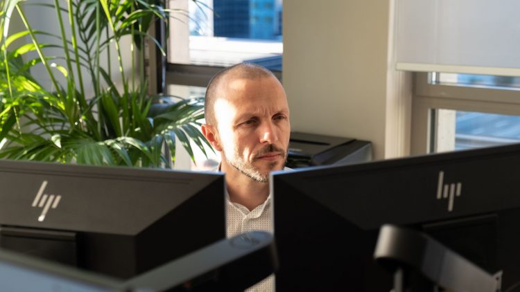 L'image montre un homme assis à un bureau, concentré sur deux écrans d'ordinateur devant lui. Il a les cheveux courts et une barbe bien taillée. Il porte une chemise blanche à motifs discrets. À l'arrière-plan, on peut voir une plante verte qui ajoute une touche de nature à l'environnement de travail. La lumière naturelle entre par une fenêtre, illuminant la scène.