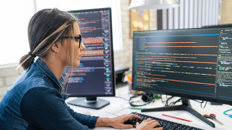 GettyImages - A woman is working on a computer, surrounded by two screens displaying code. The office environment is bright and modern, with stationery items visible on the desk.