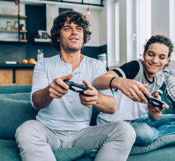 Two individuals sitting on a couch, playing video games. One is holding a game controller, focused on the screen, while the other appears to be giving advice or sharing strategies. The setting is modern and bright, with a kitchen visible in the background.