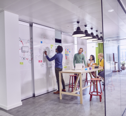 In a bright, modern office, a group of four people is engaged in a brainstorming session. A man is writing on a whiteboard, while three others, seated around a wooden table, are discussing and looking at a laptop. Colorful sticky notes are visible on the board. The space is decorated with colorful curtains and hanging lamps, creating a dynamic and collaborative atmosphere.