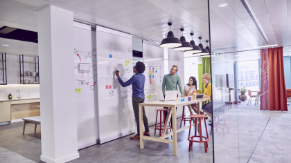 In a bright, modern office, a group of four people is engaged in a brainstorming session. A man is writing on a whiteboard, while three others, seated around a wooden table, are discussing and looking at a laptop. Colorful sticky notes are visible on the board. The space is decorated with colorful curtains and hanging lamps, creating a dynamic and collaborative atmosphere.