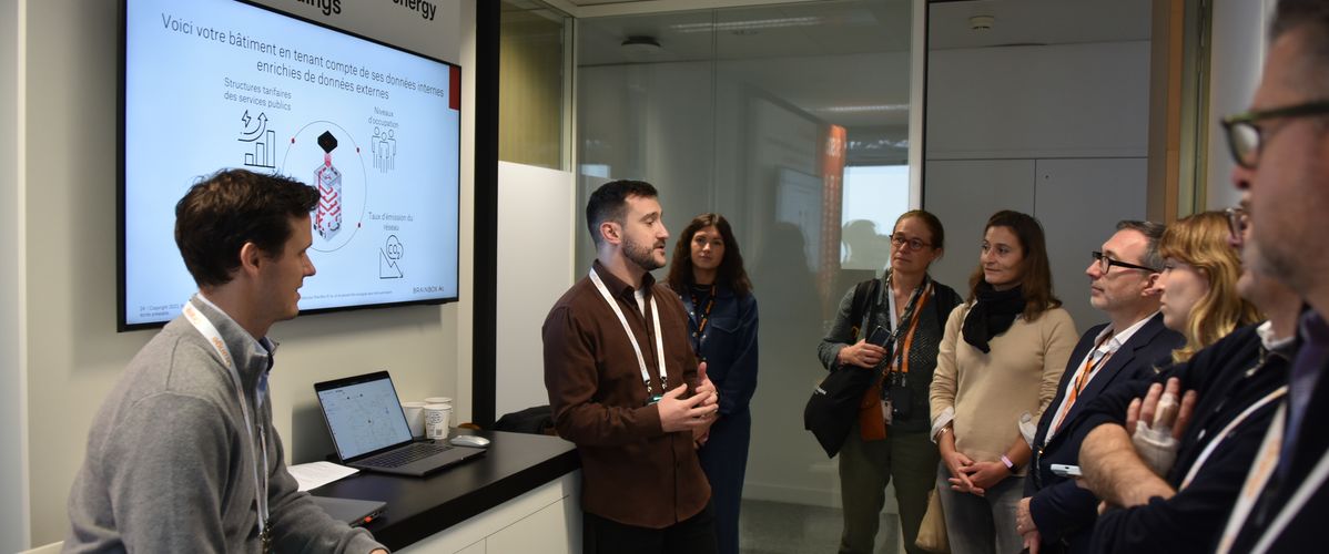 Deux présentateurs expliquent des concepts liés à BrainBox AI devant un écran affichant des graphiques et des informations. Six visiteurs se tiennent debout, attentifs.