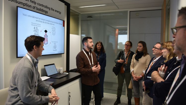 A group of people is attending a presentation of BrainBox AI at the Orange OpenTech event. Two presenter stands in front of a screen displaying graphs and information on the topic. The participants are listening attentively and appear engaged in the discussion.