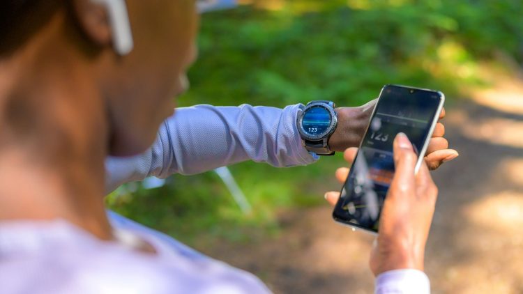 A person checks their heart rate on a smartwatch while holding a smartphone. The setting is natural, with trees in the background.