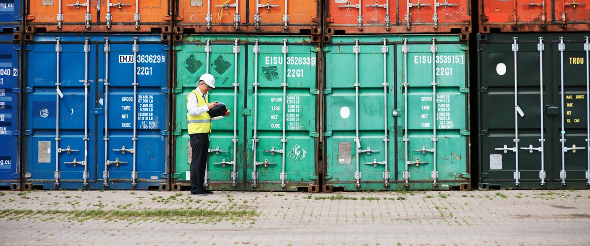 A man in a safety vest reviews documents in front of a row of colorful shipping containers at a port.