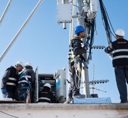 L'image montre un groupe de techniciens travaillant sur une tour de télécommunications. Ils sont engagés dans des tâches d'installation ou de maintenance, portant des casques de sécurité et des harnais, ce qui souligne l'importance de la sécurité.