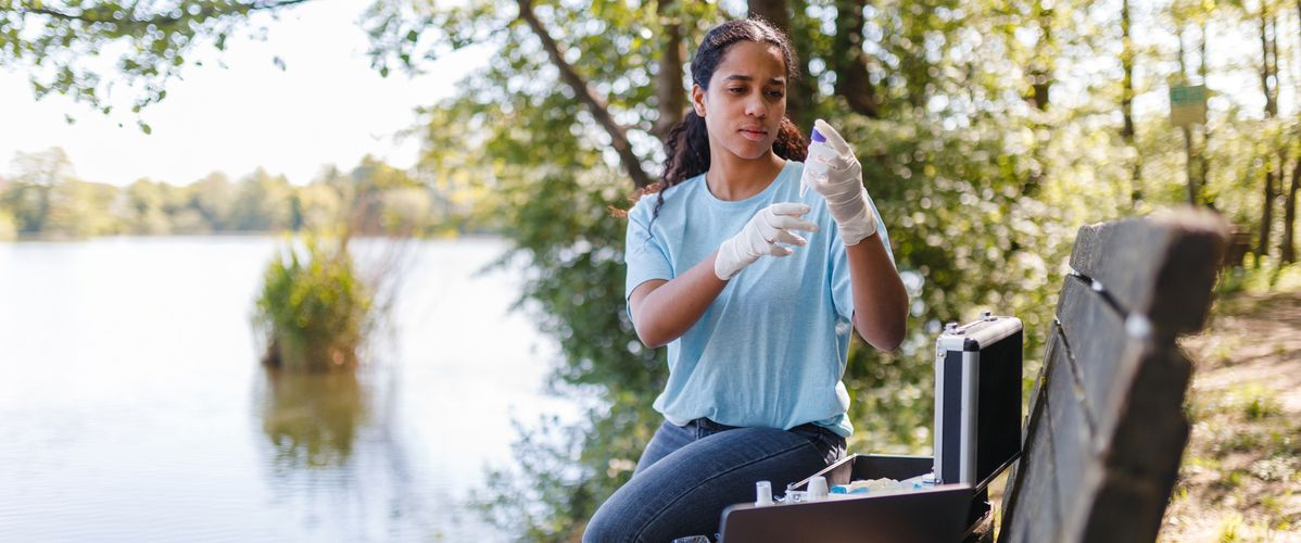 Young woman wearing gloves conducts environmental research by a lake. She uses equipment including a laptop and test kits. Trees and water in the background.