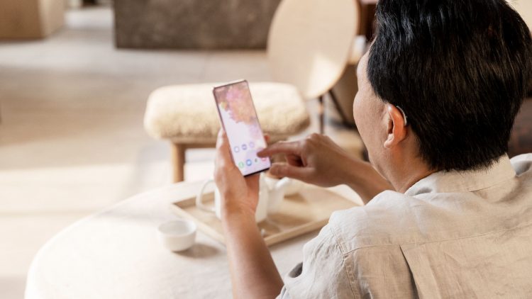 The image shows a man sitting at a table, slightly turned to the left. He is using a smartphone held in his right hand. The phone's screen displays a colorful background with app icons. On the table, there is a small white cup and a wooden tray. The man is wearing a hearing aid visible in his right ear. The environment is bright and modern, featuring furniture with soft shapes and neutral colors.