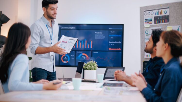 A presenter is showing graphs on a screen while colleagues listen attentively.