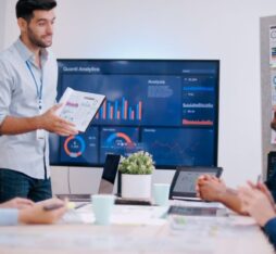 A presenter is showing graphs on a screen while colleagues listen attentively.