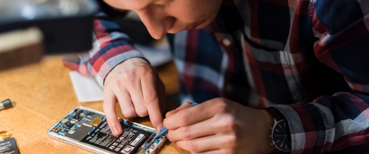 Un individu travaille sur l'intérieur d'un téléphone, utilisant un outil pour manipuler des composants électroniques. Une table en bois est visible, avec des pièces de rechange à côté.