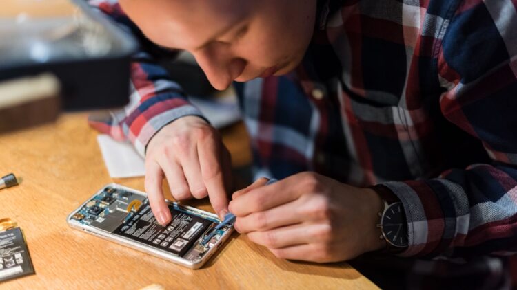 Un individu travaille sur l'intérieur d'un téléphone, utilisant un outil pour manipuler des composants électroniques. Une table en bois est visible, avec des pièces de rechange à côté.
