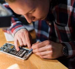 Un individu travaille sur l'intérieur d'un téléphone, utilisant un outil pour manipuler des composants électroniques. Une table en bois est visible, avec des pièces de rechange à côté.