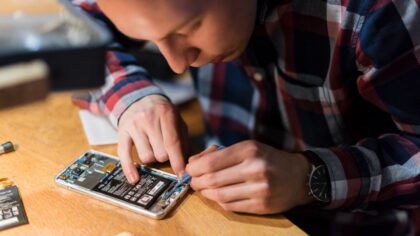 Un individu travaille sur l'intérieur d'un téléphone, utilisant un outil pour manipuler des composants électroniques. Une table en bois est visible, avec des pièces de rechange à côté.