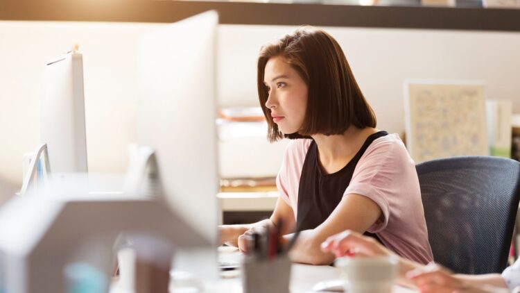 A young woman with a straight brown bob sits in an office chair, concentrating on a large desktop monitor. Natural light floods the workspace, and blurred office supplies in the foreground hint at a modern, collaborative office environment.