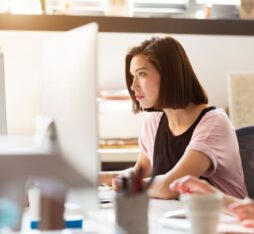 Une jeune femme, cheveux bruns coupés au carré, est assise sur une chaise de bureau et fixe attentivement un grand écran d’ordinateur. L’espace de travail baigné de lumière naturelle comporte, au premier plan, des fournitures floues qui suggèrent un bureau moderne et collaboratif.