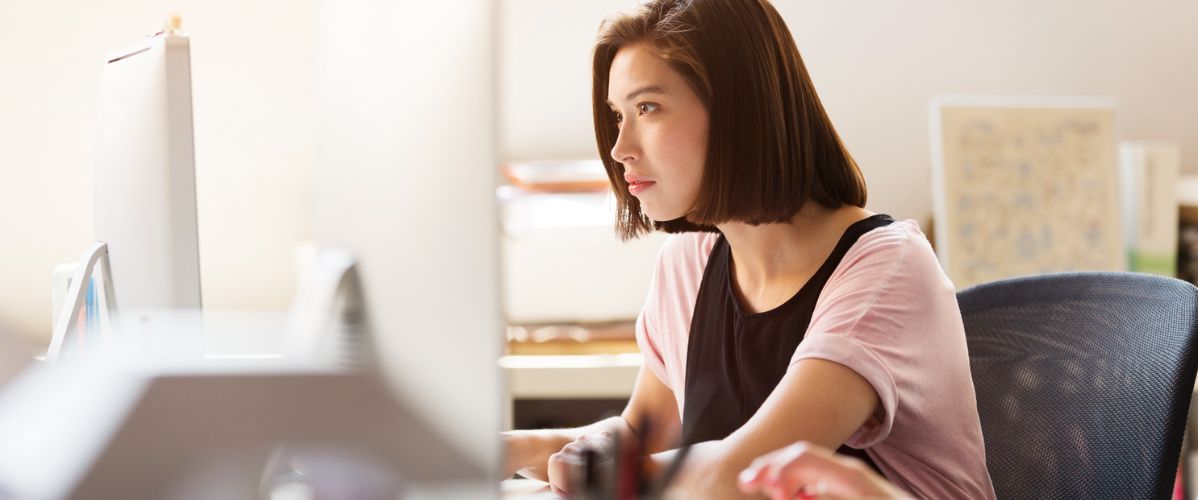 A young woman with a straight brown bob sits in an office chair, concentrating on a large desktop monitor. Natural light floods the workspace, and blurred office supplies in the foreground hint at a modern, collaborative office environment.