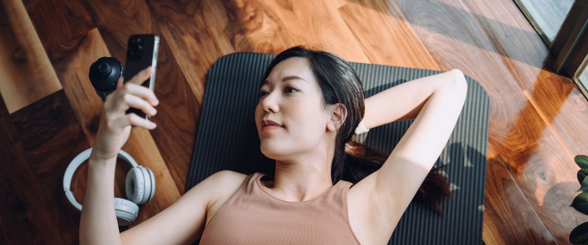 Une femme allongée sur un tapis de yoga, regardant son téléphone avec des écouteurs à côté d'elle. La lumière naturelle éclaire la pièce en bois.