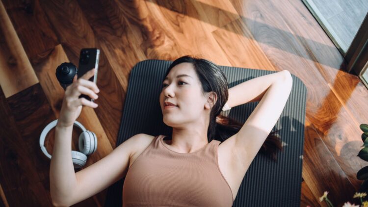 Une femme allongée sur un tapis de yoga, regardant son téléphone avec des écouteurs à côté d'elle. La lumière naturelle éclaire la pièce en bois.