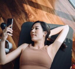 A woman lying on a yoga mat, looking at her phone with headphones beside her. Natural light illuminates the wooden room.