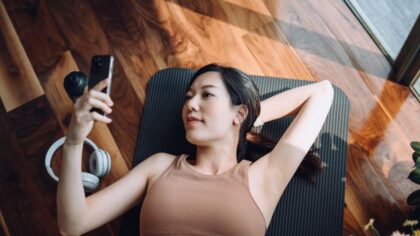 A woman lying on a yoga mat, looking at her phone with headphones beside her. Natural light illuminates the wooden room.