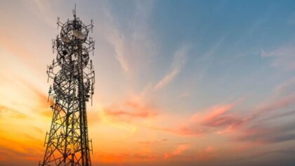 A metal telecommunications tower with multiple antennas stands against a colorful sky painted with orange, pink, and blue hues during sunset.