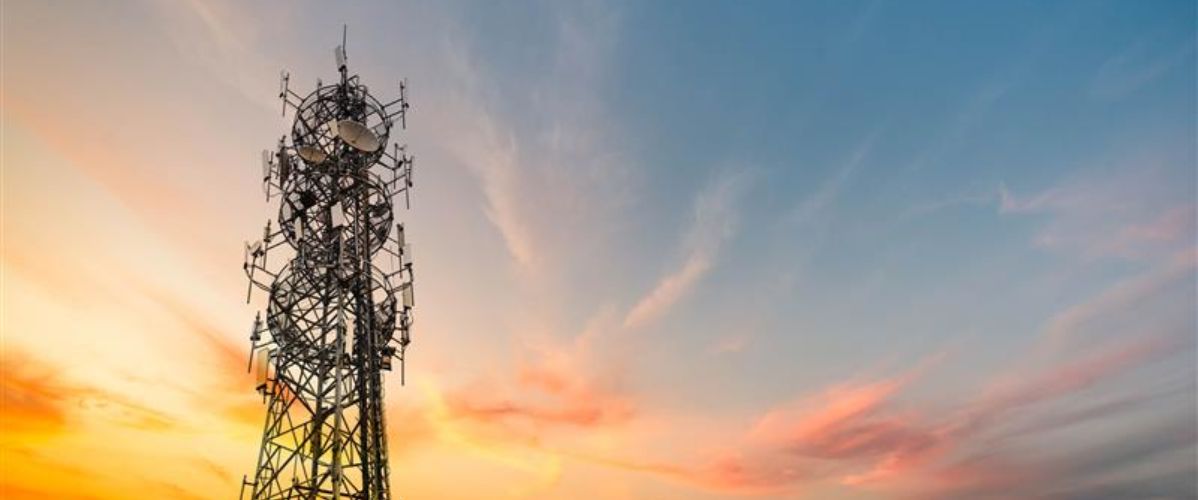 Une tour de télécommunications métallique équipée de nombreuses antennes, sur un ciel coloré par un coucher de soleil aux teintes orange, rose et bleues.