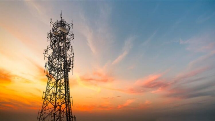 Une tour de télécommunications métallique équipée de nombreuses antennes, sur un ciel coloré par un coucher de soleil aux teintes orange, rose et bleues.