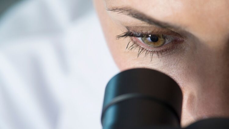 Gros plan sur le visage d’une femme en blouse blanche fixant attentivement à travers l’oculaire d’un microscope noir, avec son œil droit parfaitement aligné.