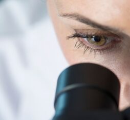 Gros plan sur le visage d’une femme en blouse blanche fixant attentivement à travers l’oculaire d’un microscope noir, avec son œil droit parfaitement aligné.