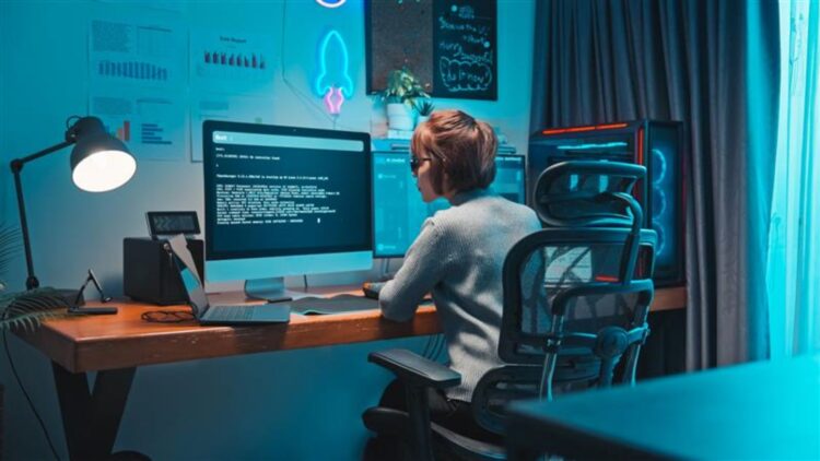 A woman sits at a desk in front of a large computer screen displaying technical text. She is wearing glasses and a light sweater. Next to her are a laptop, a desk lamp, and a computer tower. The wall behind her has charts and a neon light.