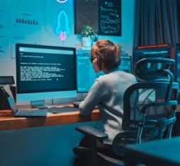 A woman sits at a desk in front of a large computer screen displaying technical text. She is wearing glasses and a light sweater. Next to her are a laptop, a desk lamp, and a computer tower. The wall behind her has charts and a neon light.