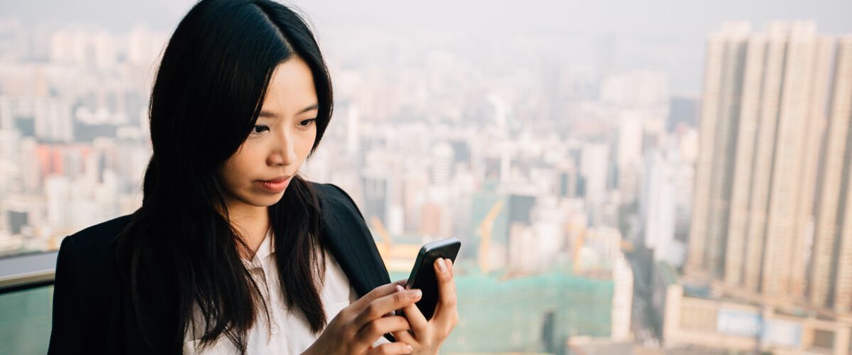 Une femme d’affaires se tient sur un balcon surplombant une grande ville. Elle porte une chemise blanche et une veste noire, et regarde attentivement l’écran de son smartphone.
