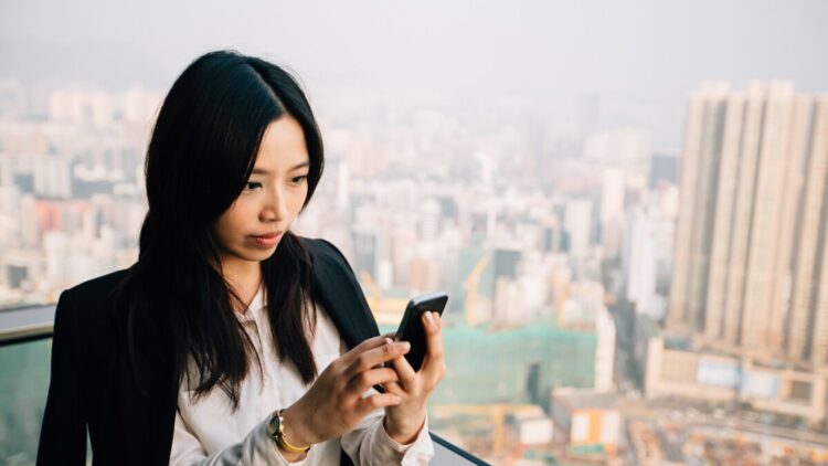 A businesswoman stands on a balcony overlooking a large city. She wears a white shirt and a black blazer, looking intently at her smartphone screen.