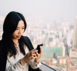 Une femme d’affaires se tient sur un balcon surplombant une grande ville. Elle porte une chemise blanche et une veste noire, et regarde attentivement l’écran de son smartphone.