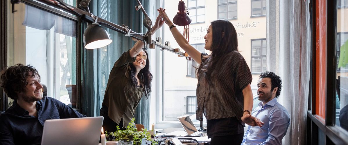Four colleagues working together in a bright office. Two women stand up and high-five in celebration, while two men sit around a table with laptops.