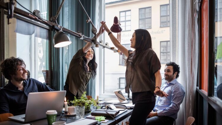 Four colleagues working together in a bright office. Two women stand up and high-five in celebration, while two men sit around a table with laptops.
