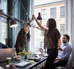 Quatre collègues travaillent ensemble dans un bureau lumineux. Deux femmes se lèvent et se tapent dans la main en signe de célébration, tandis que deux hommes sont assis autour d’une table avec des ordinateurs portables.