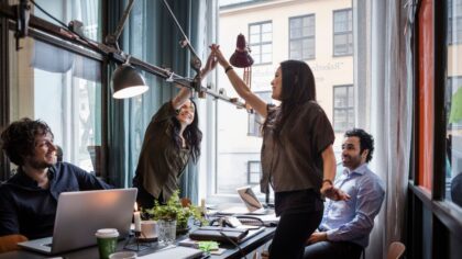 Four colleagues working together in a bright office. Two women stand up and high-five in celebration, while two men sit around a table with laptops.