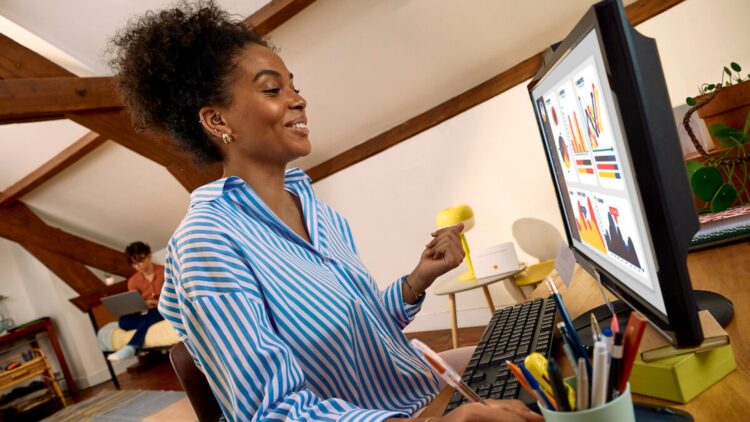 Une femme souriante est assise à un bureau dans un espace de télétravail aménagé sous les combles. Elle travaille sur un ordinateur de bureau affichant des graphiques et des tableaux de données colorés. Elle tient un stylo et prend des notes, tandis qu’une autre personne travaille en arrière-plan dans la même pièce.