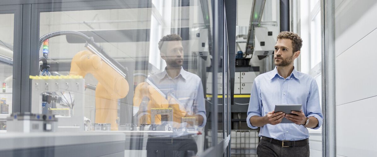 Un ingénieur tient une tablette et observe un bras robotisé industriel derrière une paroi vitrée dans un environnement de laboratoire ou d’usine automatisée.