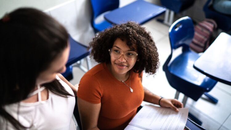 Two young women are sitting in a classroom. One of them is holding an open document on her lap while they look at each other and talk. Blue school desks and chairs are visible around them.