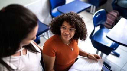 Deux jeunes femmes sont assises dans une salle de classe. L’une tient un document ouvert sur ses genoux tandis qu’elles échangent un regard et discutent. Des tables et des chaises d’école bleues sont visibles autour d’elles.