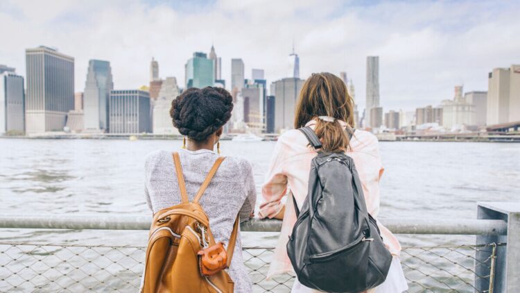 Deux femmes portant des sacs à dos regardent l’horizon depuis une promenade au bord de l’eau, face à une grande ville et ses gratte-ciel.