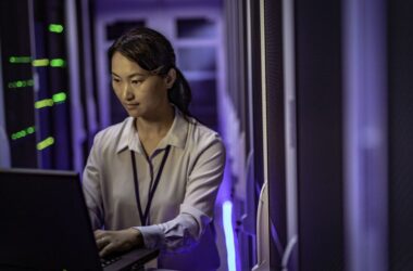 Une femme travaille sur un ordinateur portable au sein d’un data center. Elle se tient entre des rangées de serveurs informatiques éclairés par des voyants lumineux bleus et verts, dans un environnement technique et sécurisé.