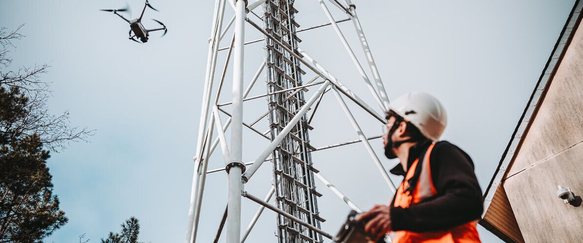 Un technicien portant un casque et un gilet de sécurité pilote un drone près d’une tour de télécommunication.