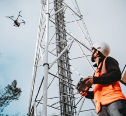 Un technicien portant un casque et un gilet de sécurité pilote un drone près d’une tour de télécommunication.