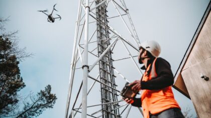 Un technicien portant un casque et un gilet de sécurité pilote un drone près d’une tour de télécommunication.